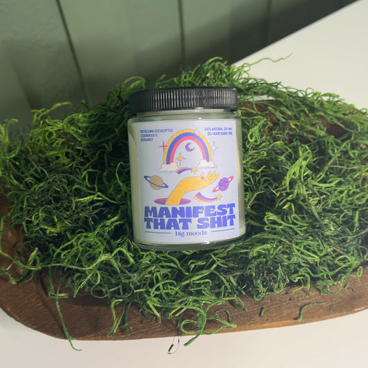 Jar labeled 'Manifest That Shit' on a bed of green moss with a wooden board underneath.