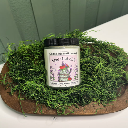 Jar of 'Sage that Shit' candle on a bed of green moss with a wooden board underneath.