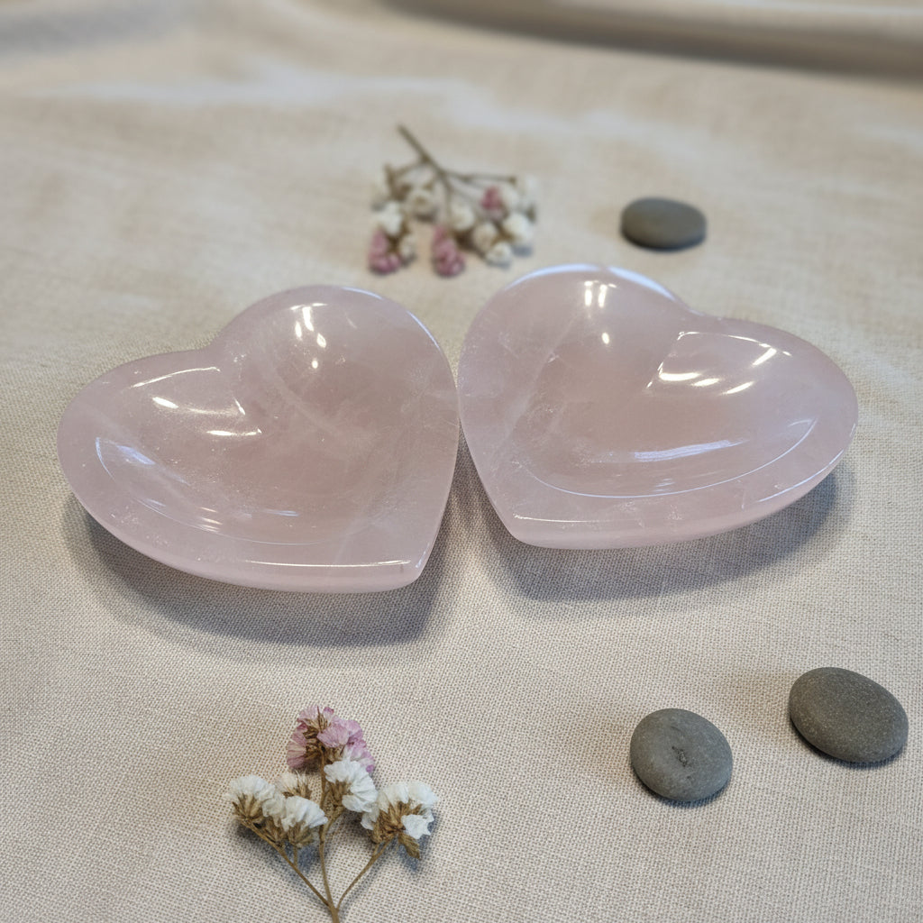 Two heart-shaped gemstone bowls on a tan surface with flowers and pebbles surrounding
