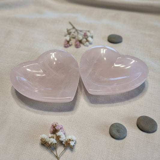 Two heart-shaped gemstone bowls on a tan surface with flowers and pebbles surrounding
